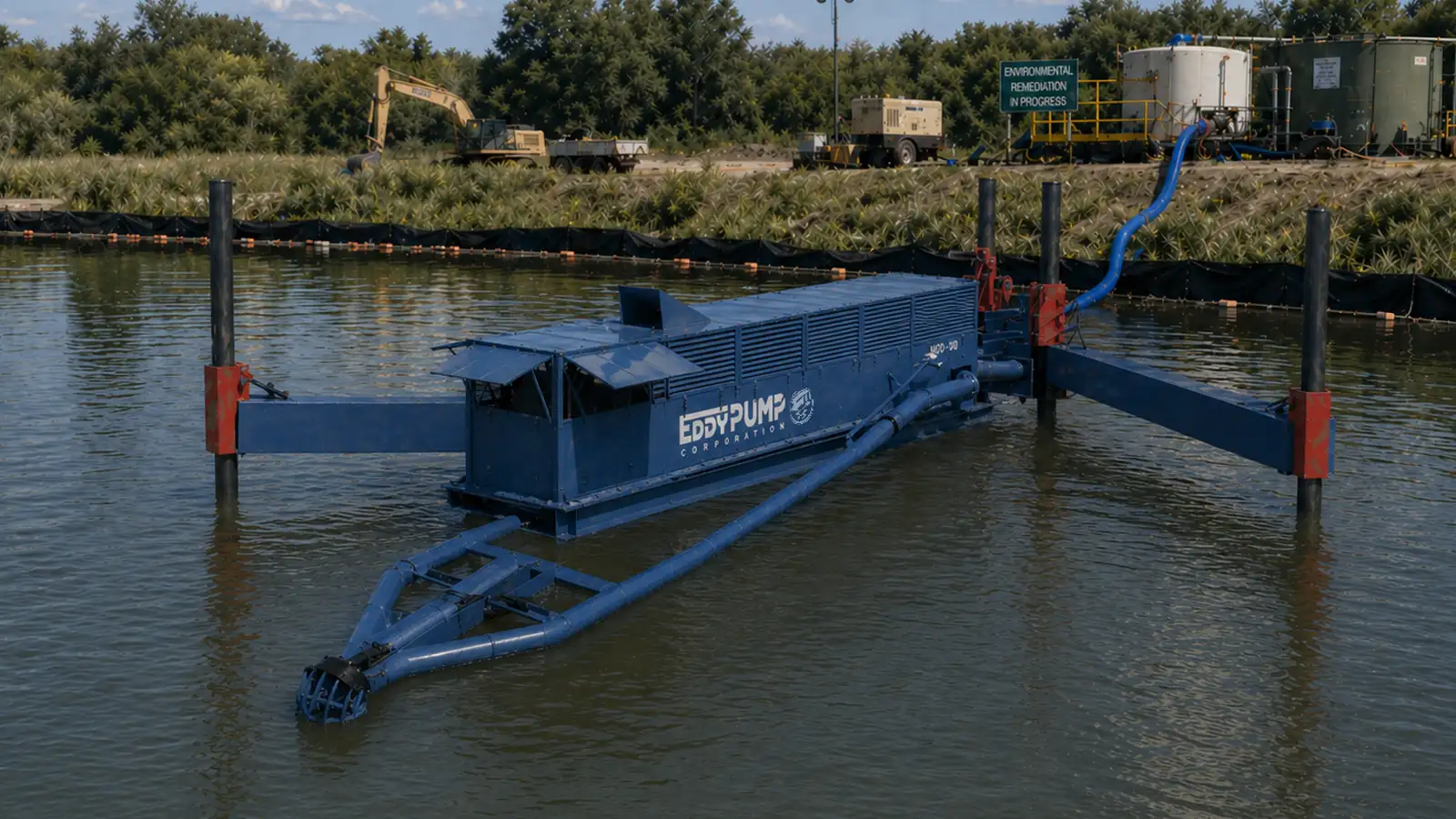 Large enclosed barge-style dredge unit guided between vertical steel poles in a calm water body with a blue discharge pipe extending to shore, positioned at an environmental remediation site with storage tanks and an excavator on the vegetated bank