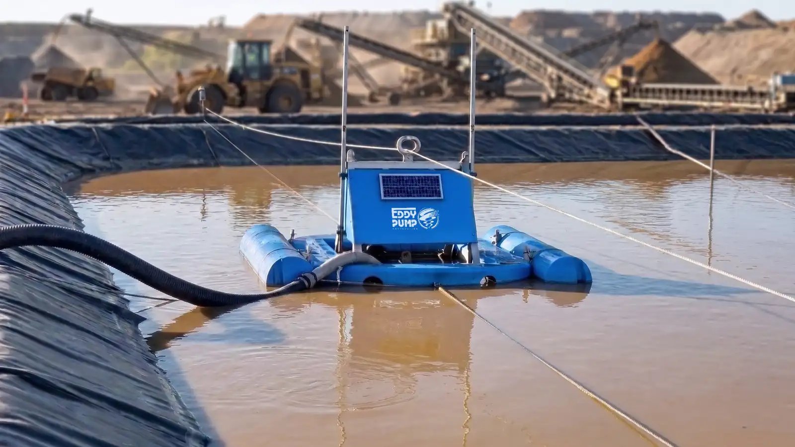 Solar-powered floating dredge on pontoons dredging a lined mine tailings pond, with haul trucks, loader and aggregate conveyors in the background.