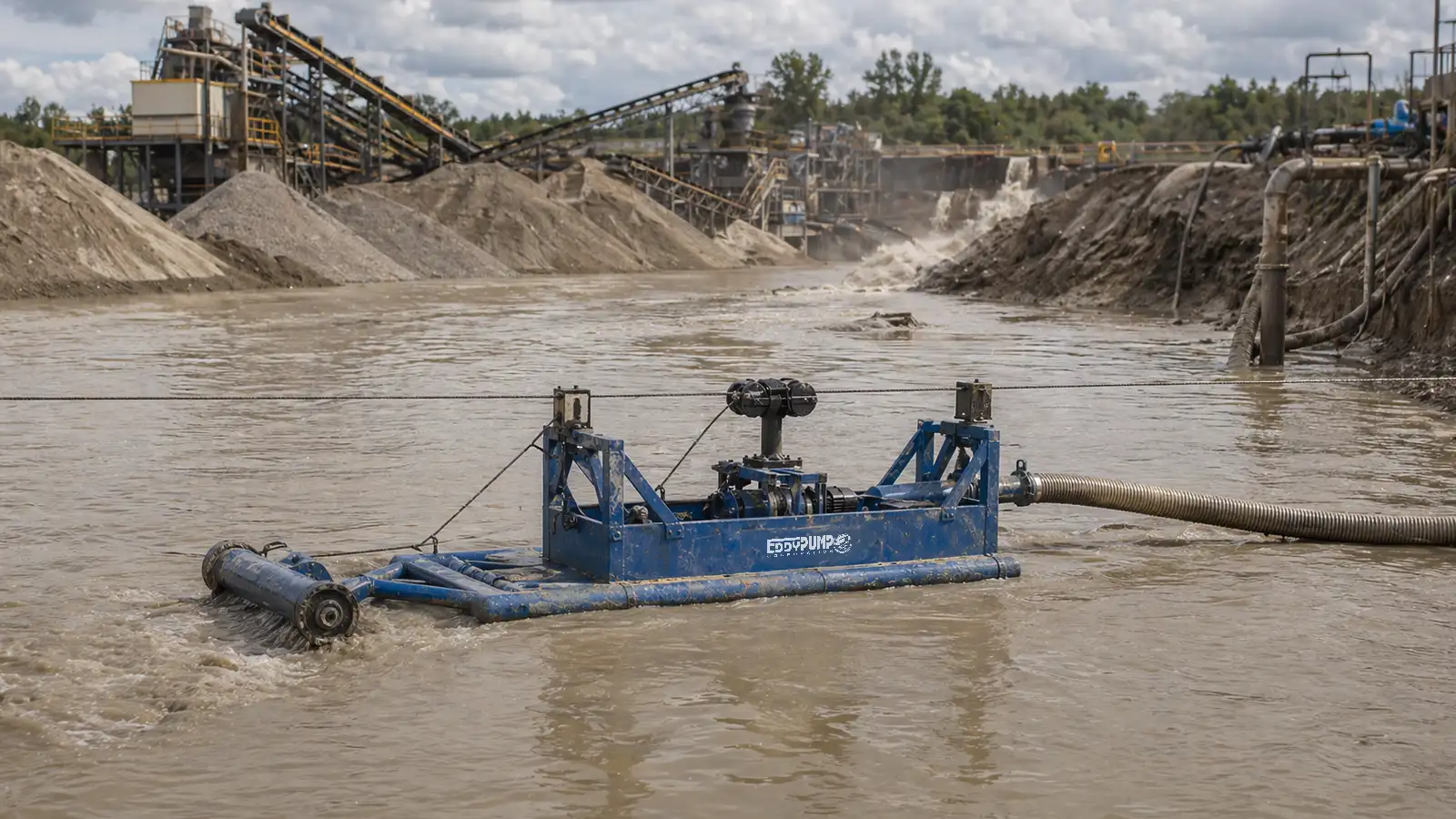 EDDY Pump floating dredge system actively pumping slurry from a turbid mine process pond with aggregate stockpiles and industrial conveyor systems in the background
